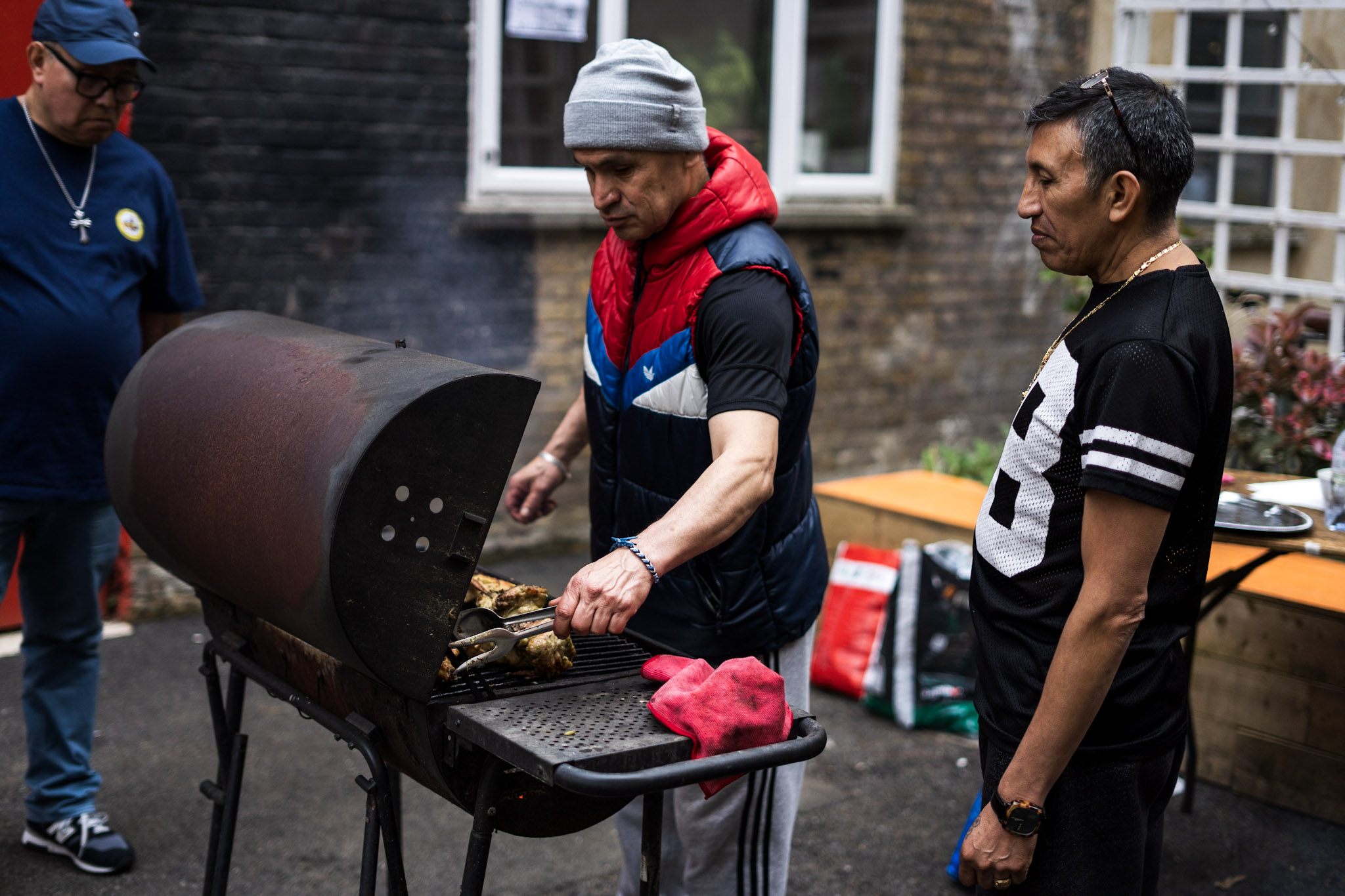 Three people are gathered around an outdoor barbecue grill. One person is tending to the meat on the barbecue. Another person, wearing a sports jersey, watches. A third person stands nearby, also watching on. A brick building is in the background.